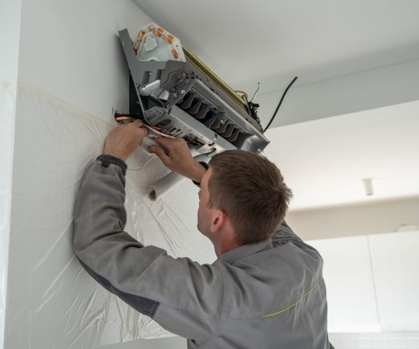 Professional man technician in uniform installing air conditioner in modern apartment on wall near window. Maintenance and repair service concept. Creating pleasant microclimate for living in flat.