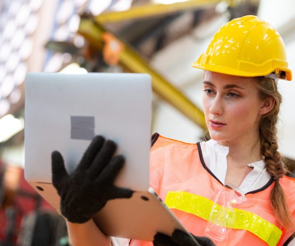 Female industrial engineer or technician worker in hard helmet and uniform using laptop checking on robotic arm machine. woman work hard in heavy technology invention industry manufacturing factory